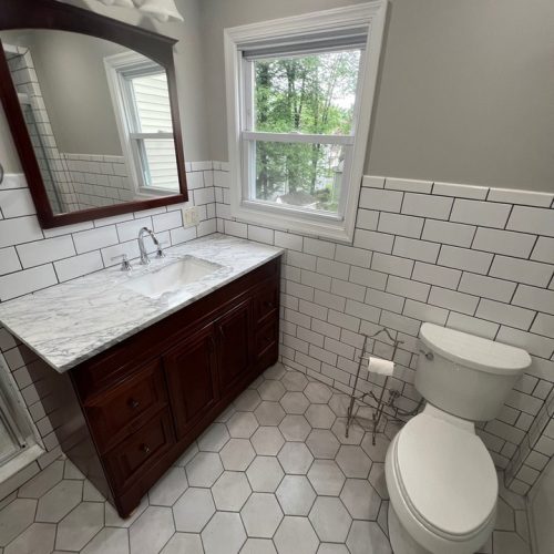 Bathroom with gray hexagonal floor tiles, a dark wood vanity with a marble countertop, white subway tile walls, a toilet, and a window with greenery outside.